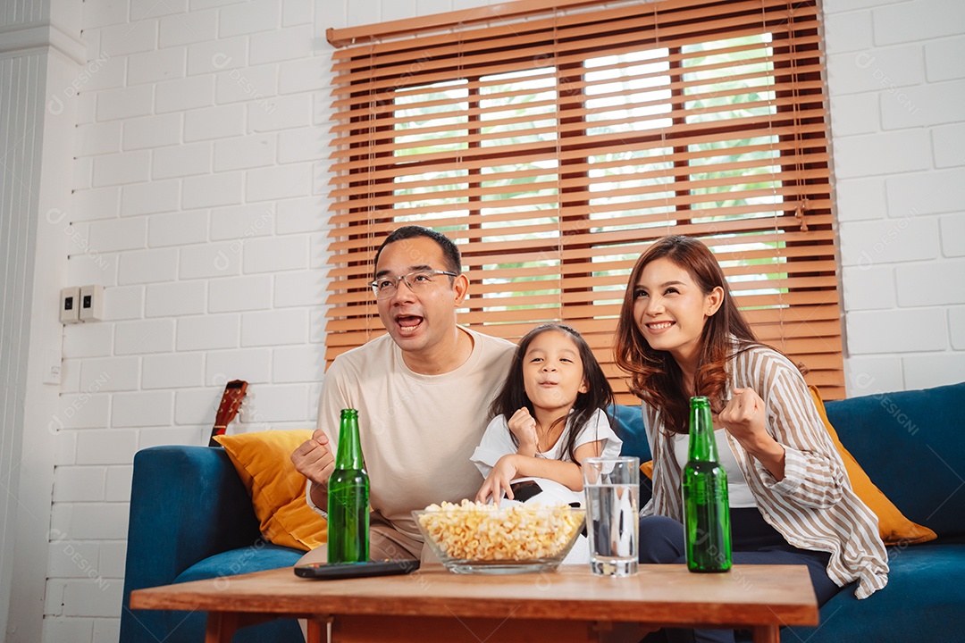 Família asiática com a filha assistindo futebol esportivo torcendo animado comendo pipoca no sofá na sala