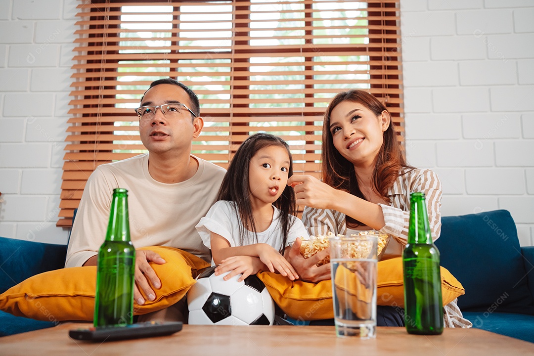 Família asiática com a filha assistindo futebol esportivo torcendo animado comendo pipoca no sofá na sala