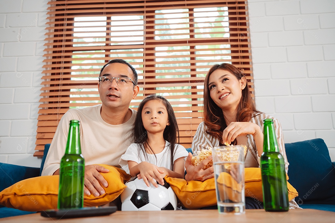 Família asiática com a filha assistindo futebol esportivo torcendo animado comendo pipoca no sofá na sala