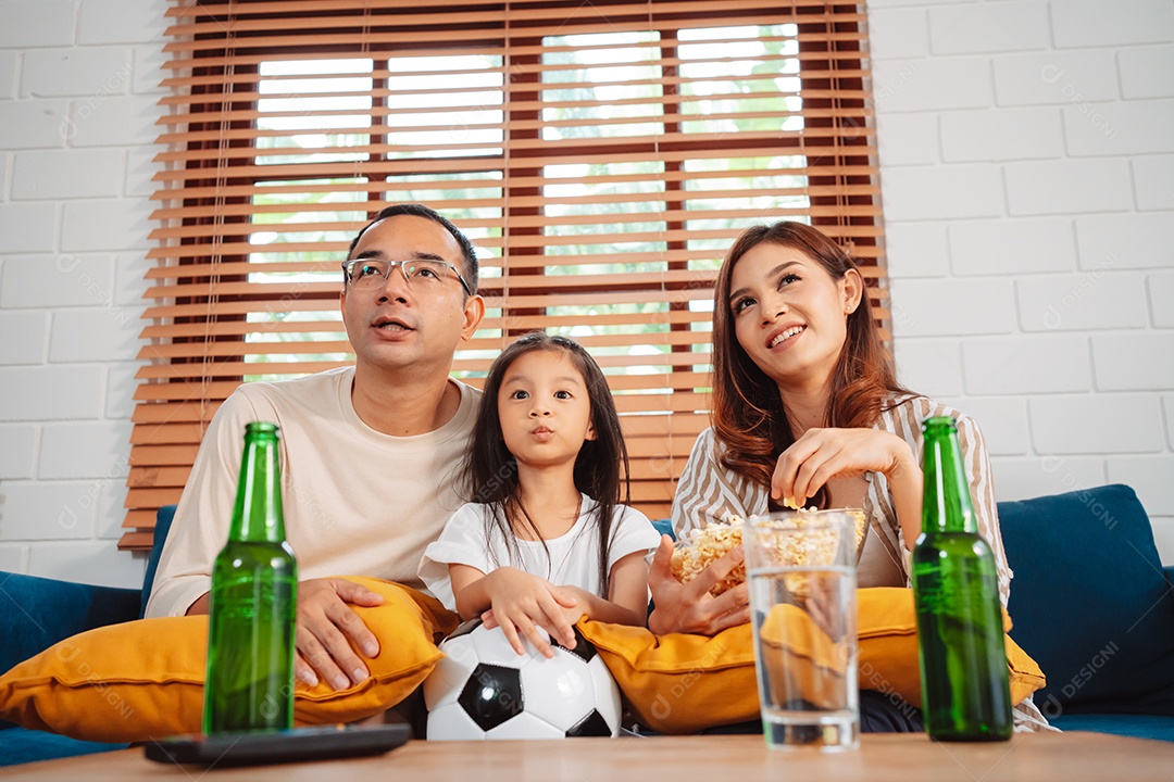 Família asiática com a filha assistindo futebol esportivo torcendo animado comendo pipoca no sofá na sala