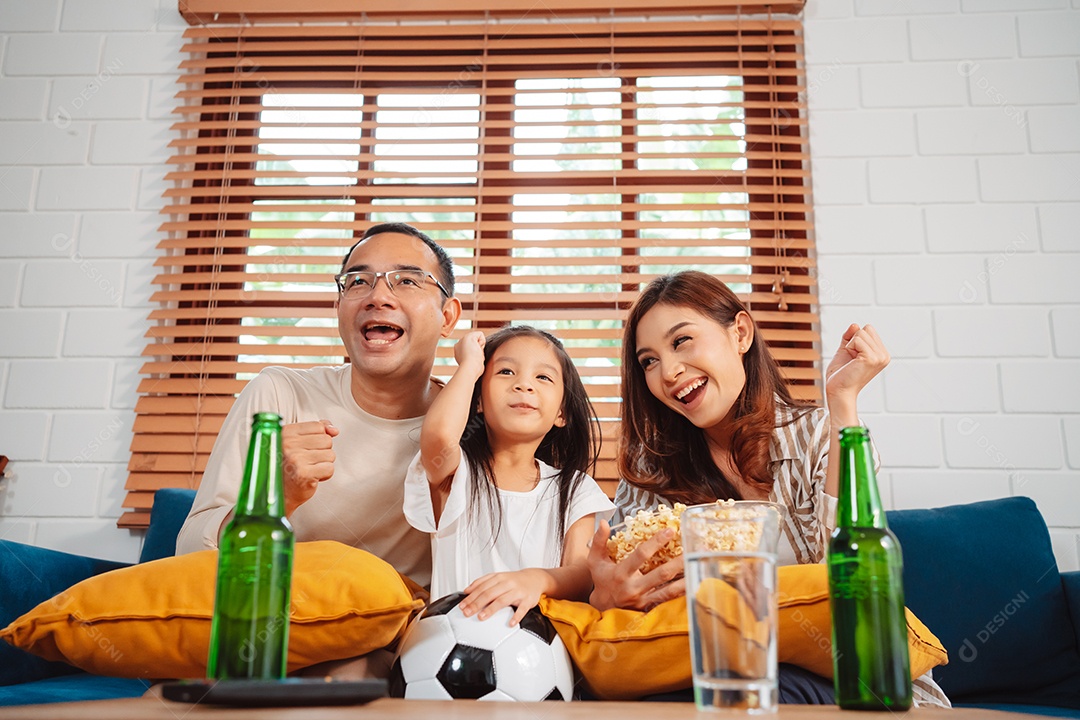 Família asiática com a filha assistindo futebol esportivo torcendo animado comendo pipoca no sofá na sala