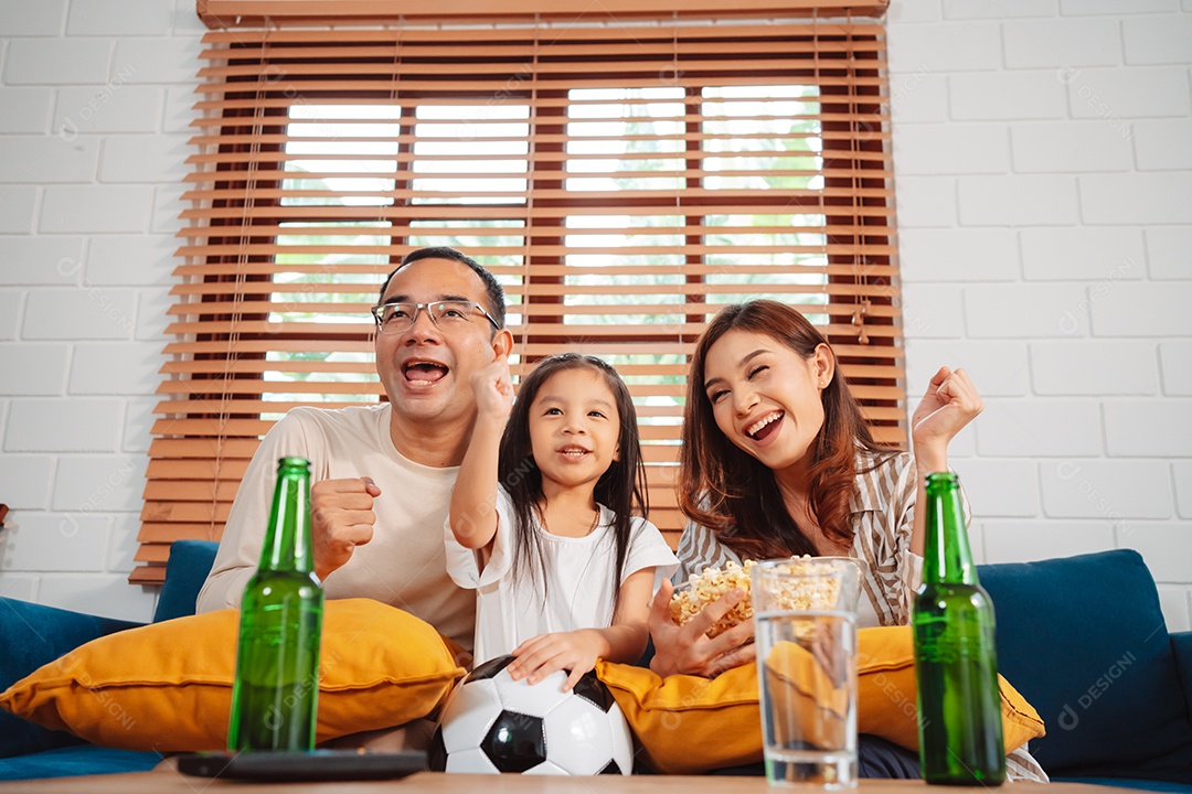 Família asiática com a filha assistindo futebol esportivo torcendo animado comendo pipoca no sofá na sala