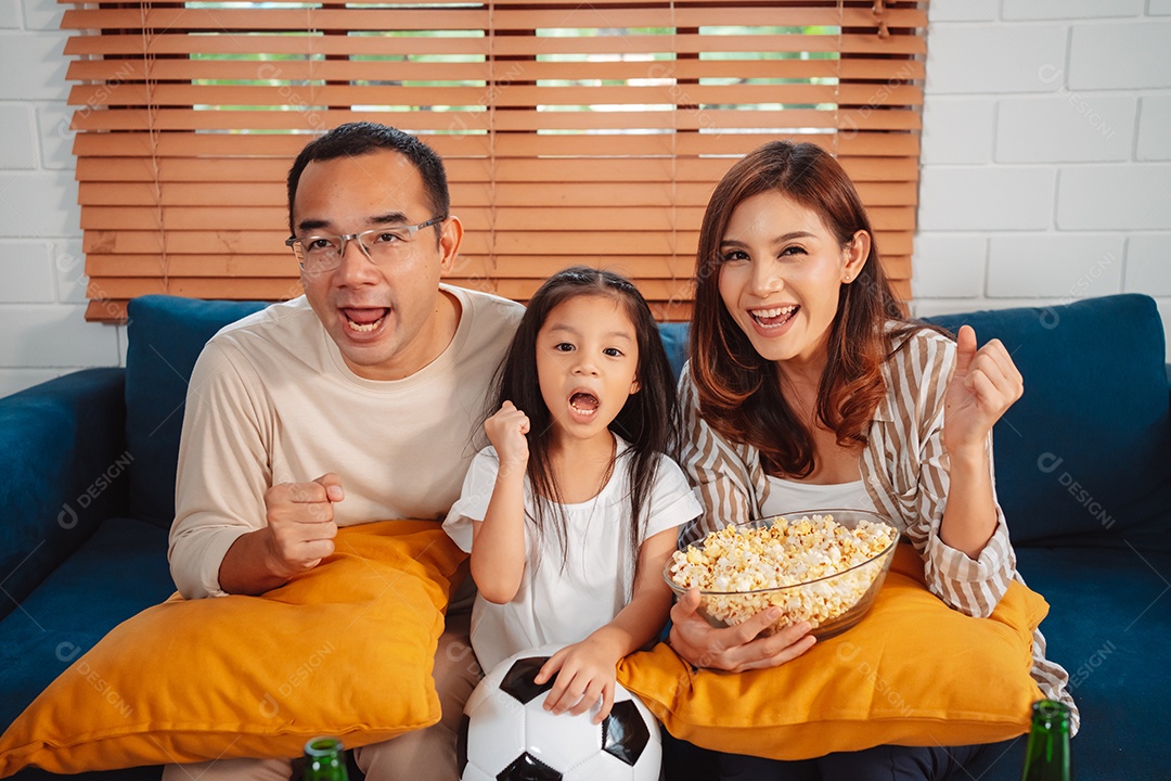 Família asiática com a filha assistindo futebol esportivo torcendo animado comendo pipoca no sofá na sala