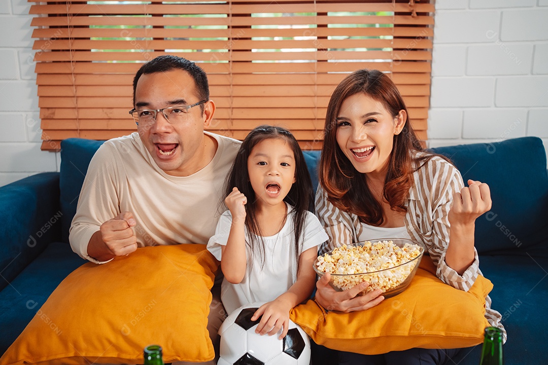 Família asiática com a filha assistindo futebol esportivo torcendo animado comendo pipoca no sofá na sala