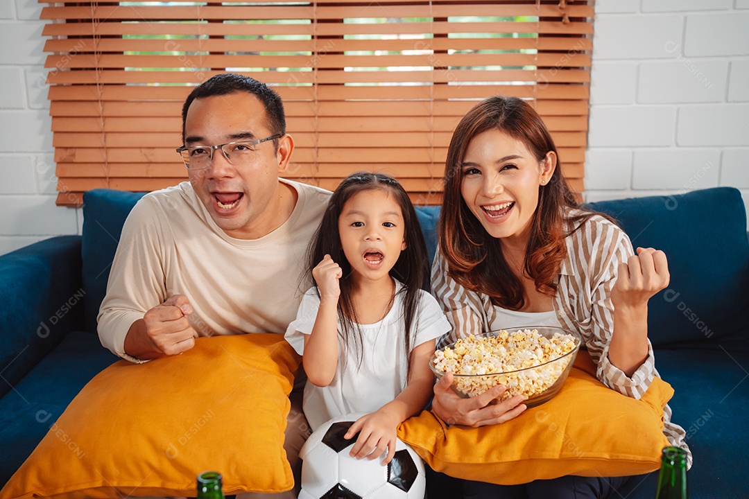 Família asiática com a filha assistindo futebol esportivo torcendo animado comendo pipoca no sofá na sala