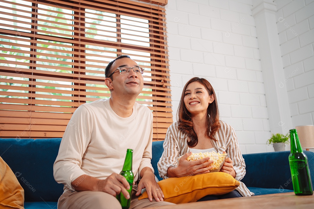 Casal asiático assistindo futebol esportivo torcendo animado no sofá na sala de estar em casa.