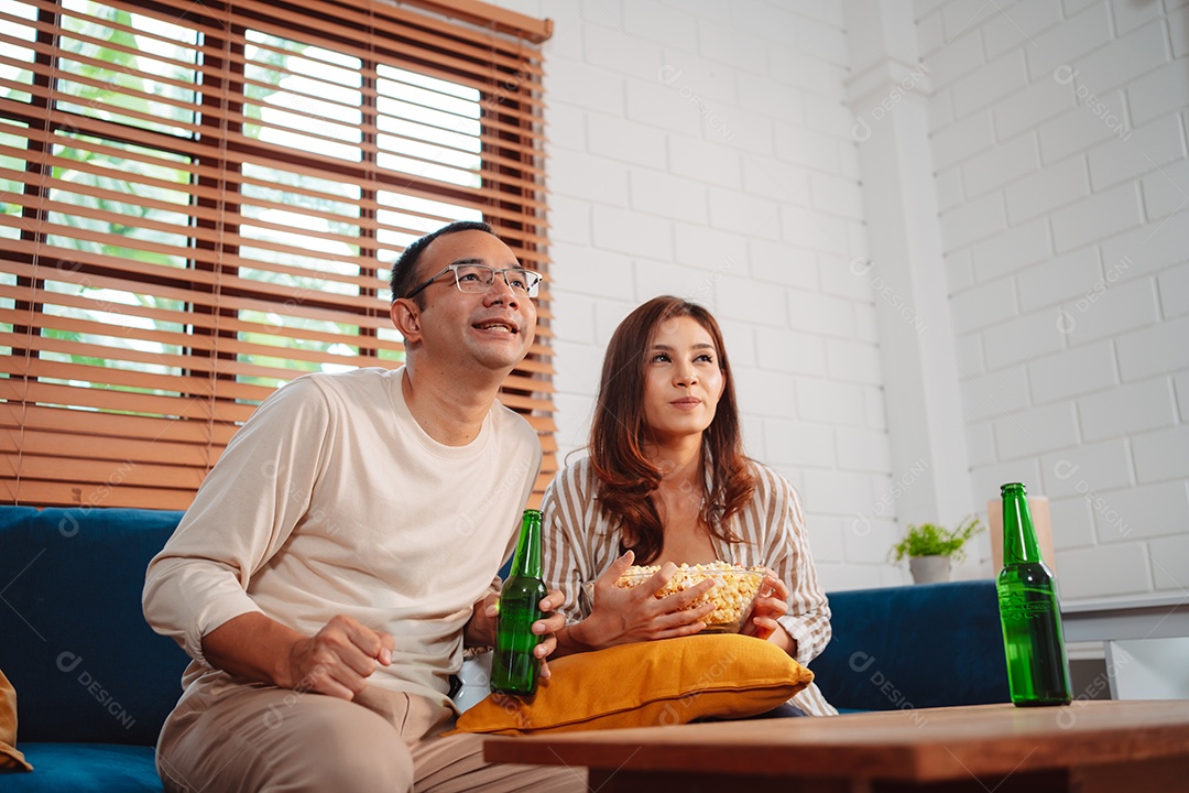 Casal asiático assistindo futebol esportivo torcendo animado no sofá na sala de estar em casa.