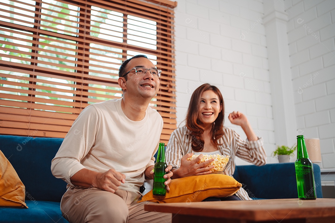 Casal asiático assistindo futebol esportivo torcendo animado no sofá na sala de estar em casa.