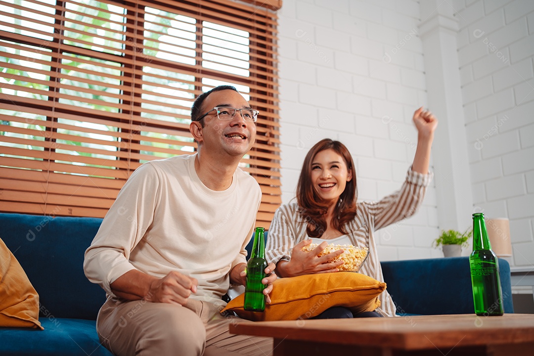 Casal asiático assistindo futebol esportivo torcendo animado no sofá na sala de estar em casa.