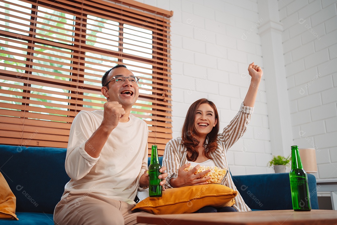 Casal asiático assistindo futebol esportivo torcendo animado no sofá na sala de estar em casa.