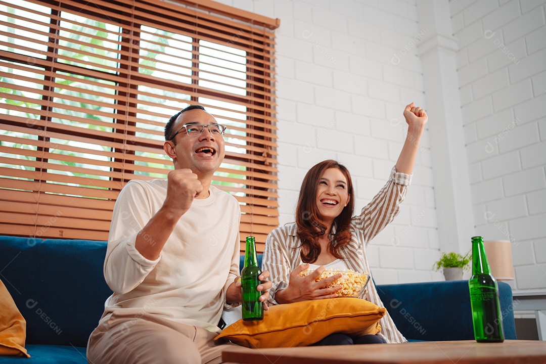 Casal asiático assistindo futebol esportivo torcendo animado no sofá na sala de estar em casa.