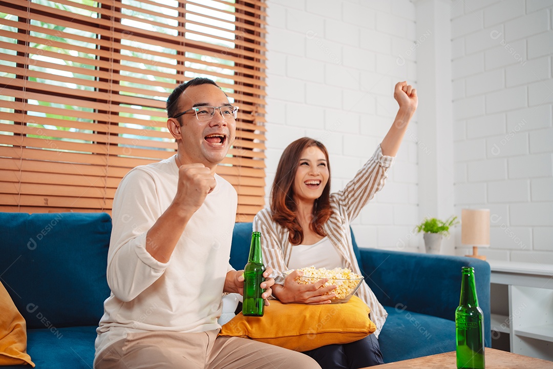 Casal asiático assistindo futebol esportivo torcendo animado no sofá na sala de estar em casa.