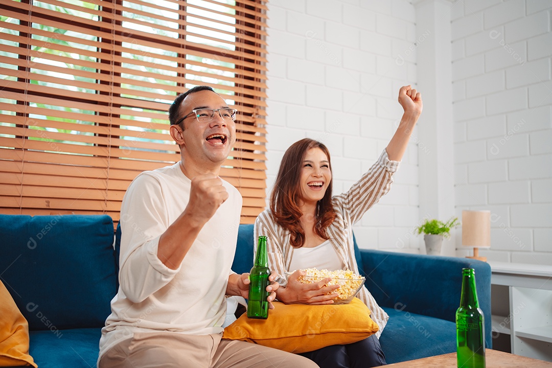 Casal asiático assistindo futebol esportivo torcendo animado no sofá na sala de estar em casa.
