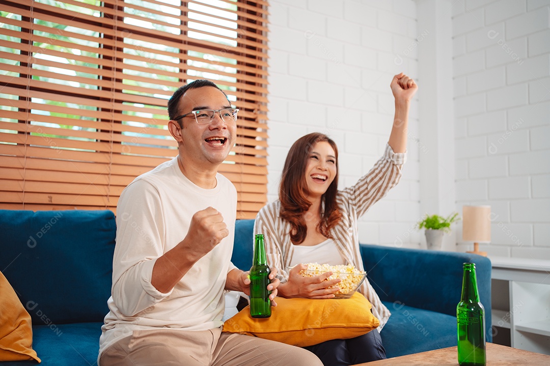 Casal asiático assistindo futebol esportivo torcendo animado no sofá na sala de estar em casa.