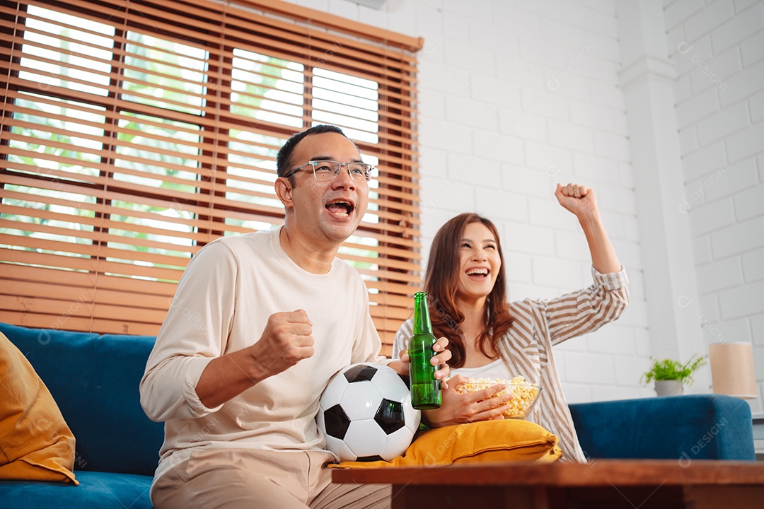 Casal asiático assistindo futebol esportivo torcendo animado no sofá na sala de estar em casa.