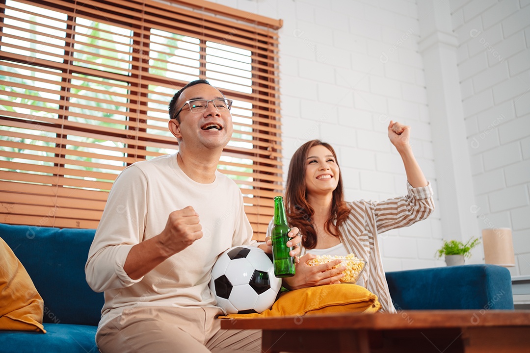 Casal asiático assistindo futebol esportivo torcendo animado no sofá na sala de estar em casa.