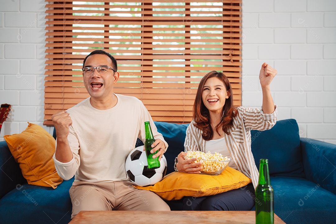 Casal asiático assistindo futebol esportivo torcendo animado no sofá na sala de estar em casa.