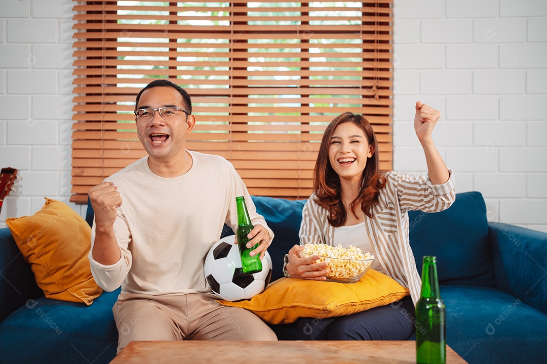 Casal asiático assistindo futebol esportivo torcendo animado no sofá na sala de estar em casa.