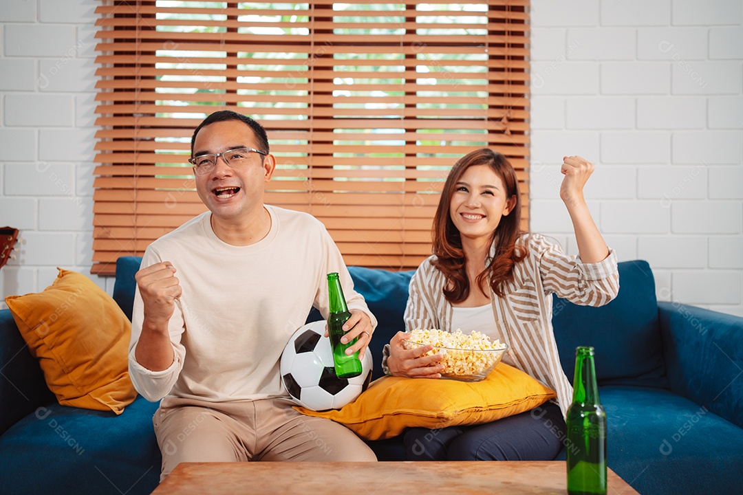 Casal asiático assistindo futebol esportivo torcendo animado no sofá na sala de estar em casa.