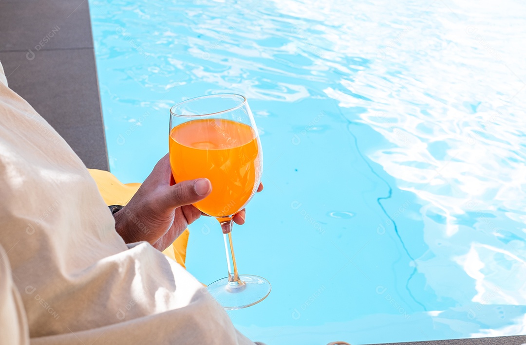 A mão do homem segurando um copo de suco de laranja à beira da piscina, lazer