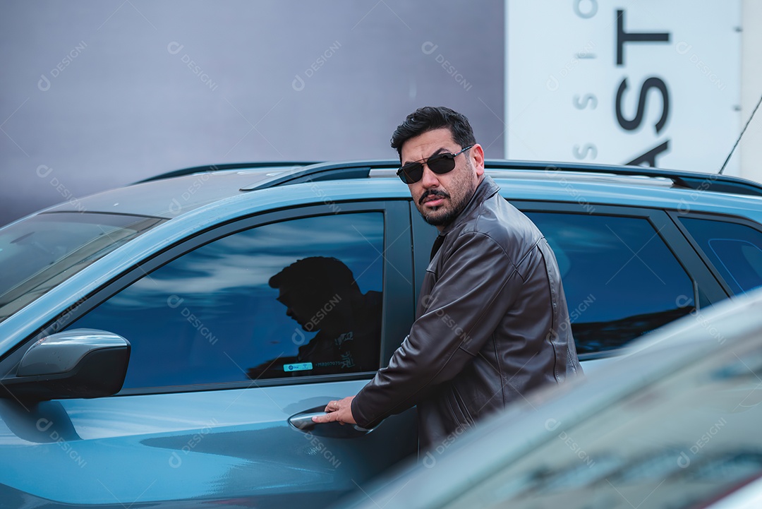 Homem lindo de meia idade serio e usando óculos de sol e jaqueta ao lado de uma carro