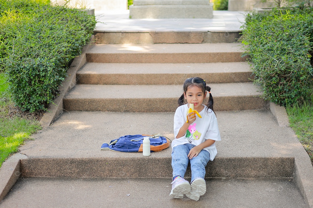 Garotinha asiática segurando e comendo banana na escola.