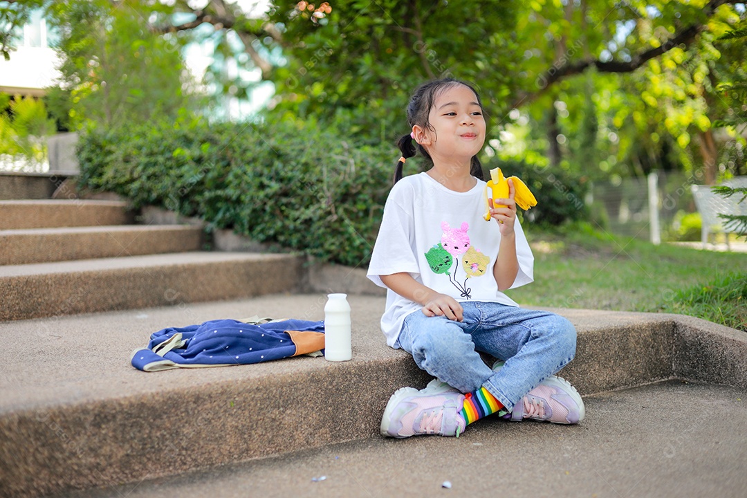 Garotinha asiática segurando e comendo banana na escola.