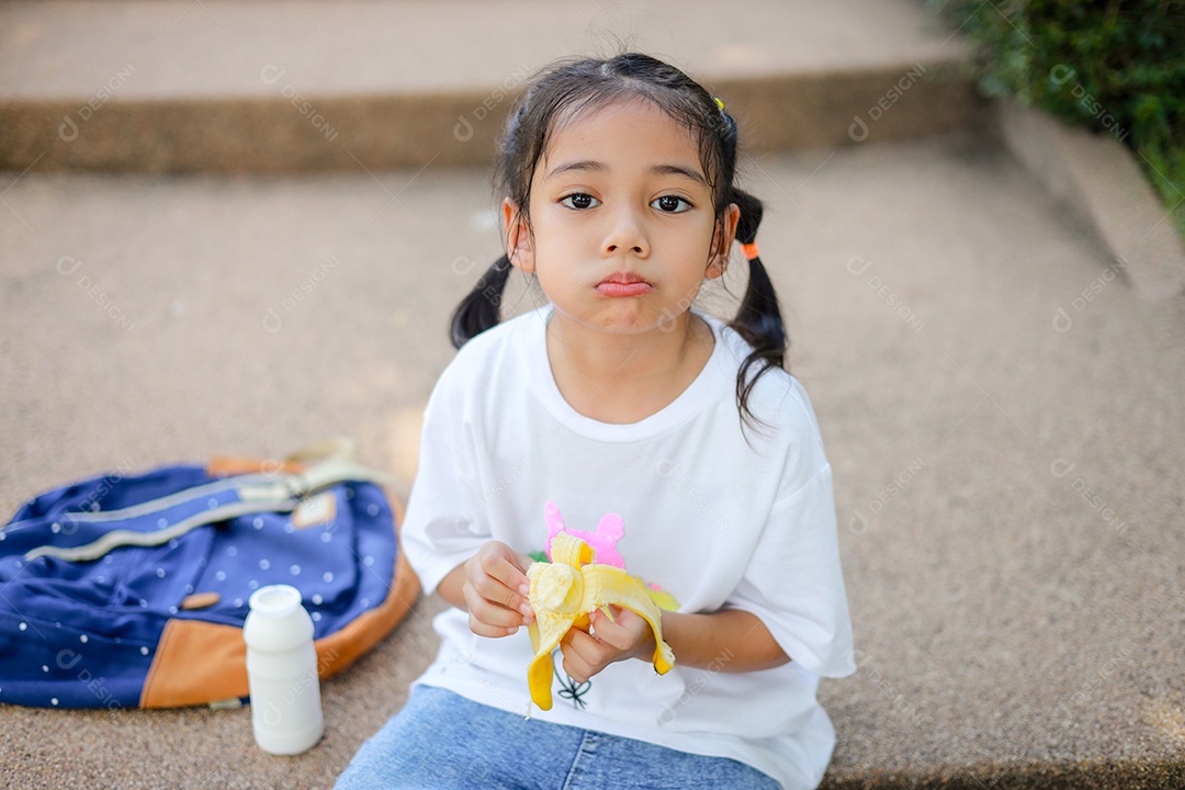 Garotinha asiática segurando e comendo banana na escola.