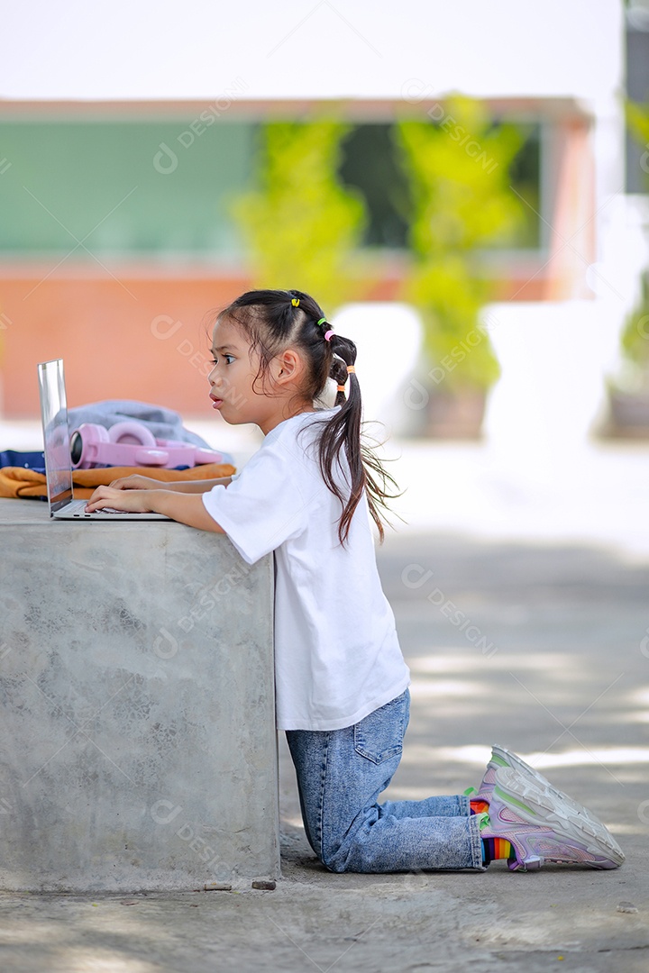 Garotinha asiática usa um laptop para aprender na escola. A criança sorri feliz e obtém conhecimento remotamente.