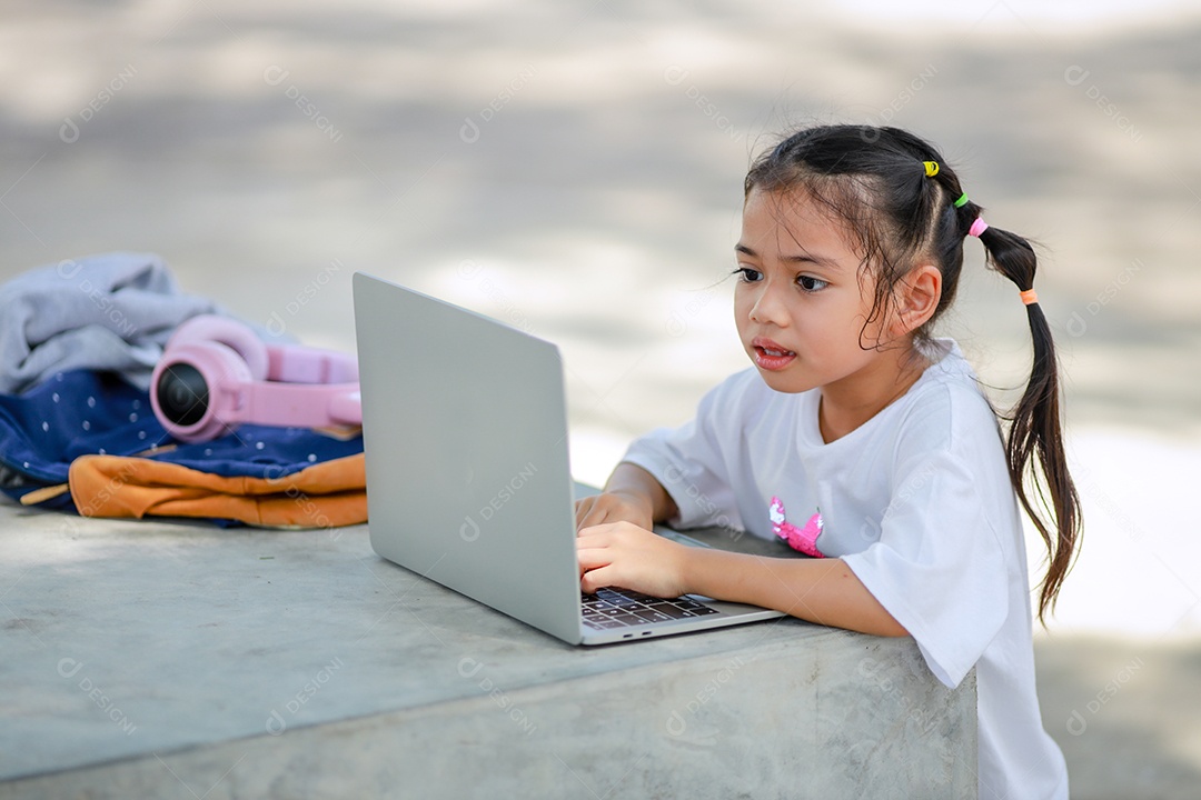 Garotinha asiática usa um laptop para aprender na escola. A criança sorri feliz e obtém conhecimento remotamente.