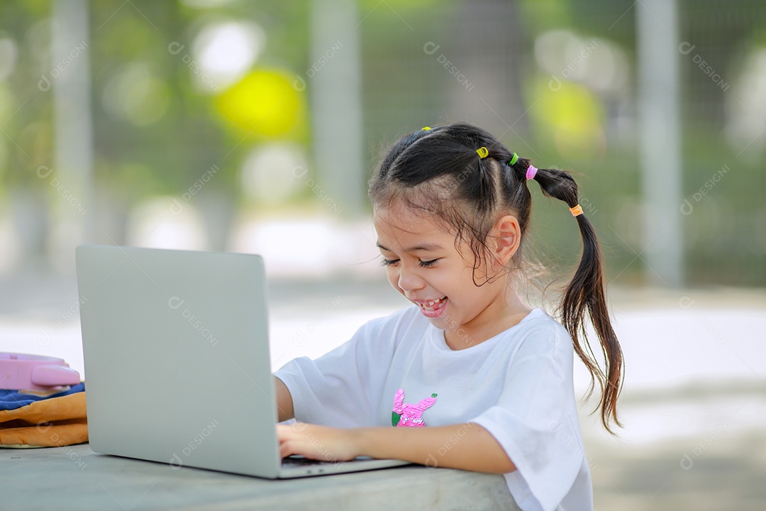 Garotinha asiática usa um laptop para aprender na escola. A criança sorri feliz e obtém conhecimento remotamente.