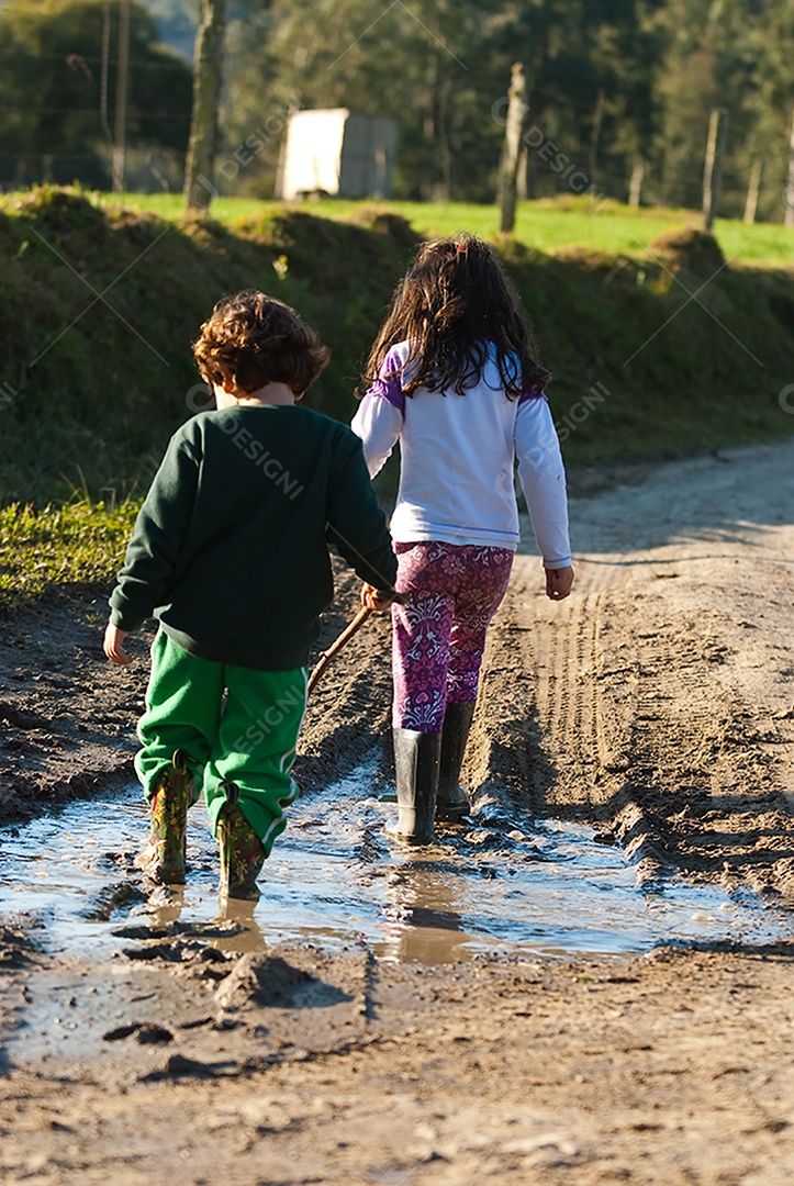 Duas crianças de bota andando sobre poça de lama