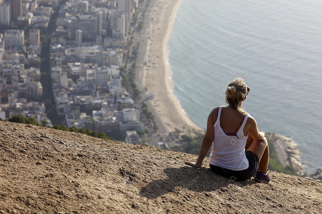 Mulher sentada no morro apreciando vista para o mar