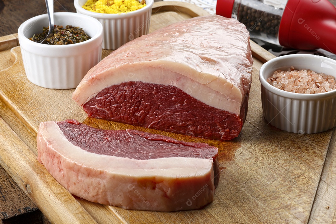 Peça de carne crua sobre tábua de madeira com temperos ao lado