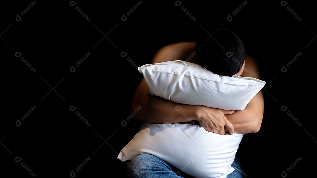 Homem de meia-idade sem camisa sentado estressado, segurando a cabeça com as mãos e abraçando o travesseiro branco sobre fundo preto.
