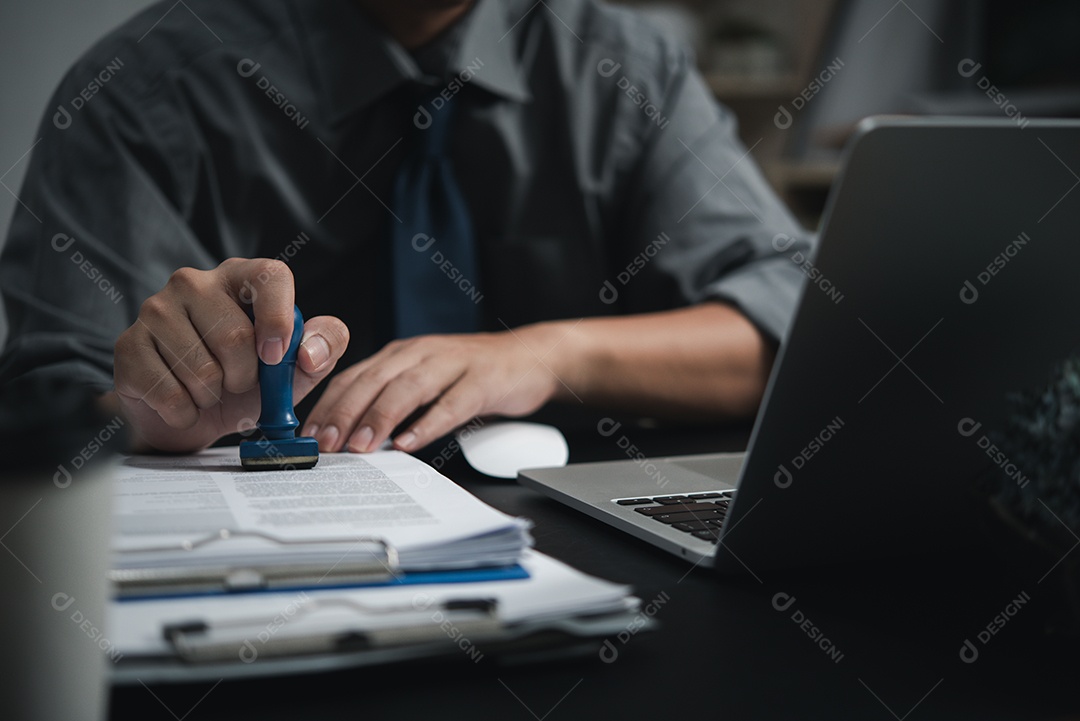 Mão de homem carimbando aprovação assinando contrato de documento ou papelada na mesa.