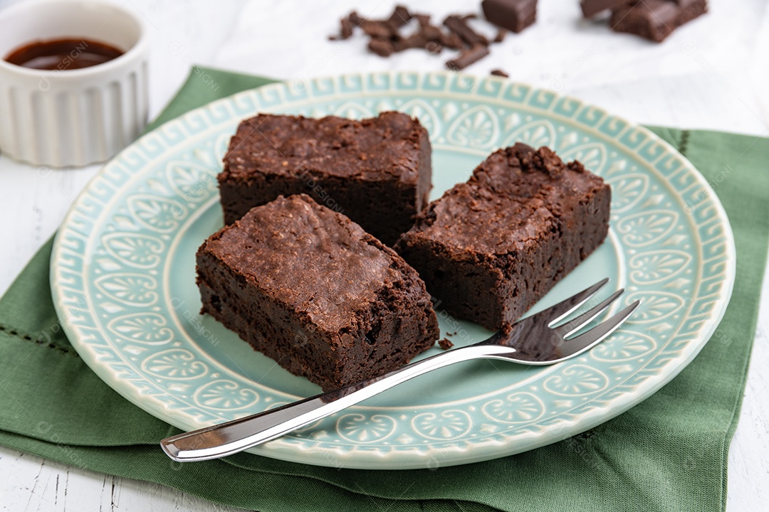 Brownies em um prato verde com pedaços de chocolate e calda sobre a mesa de madeira branca.