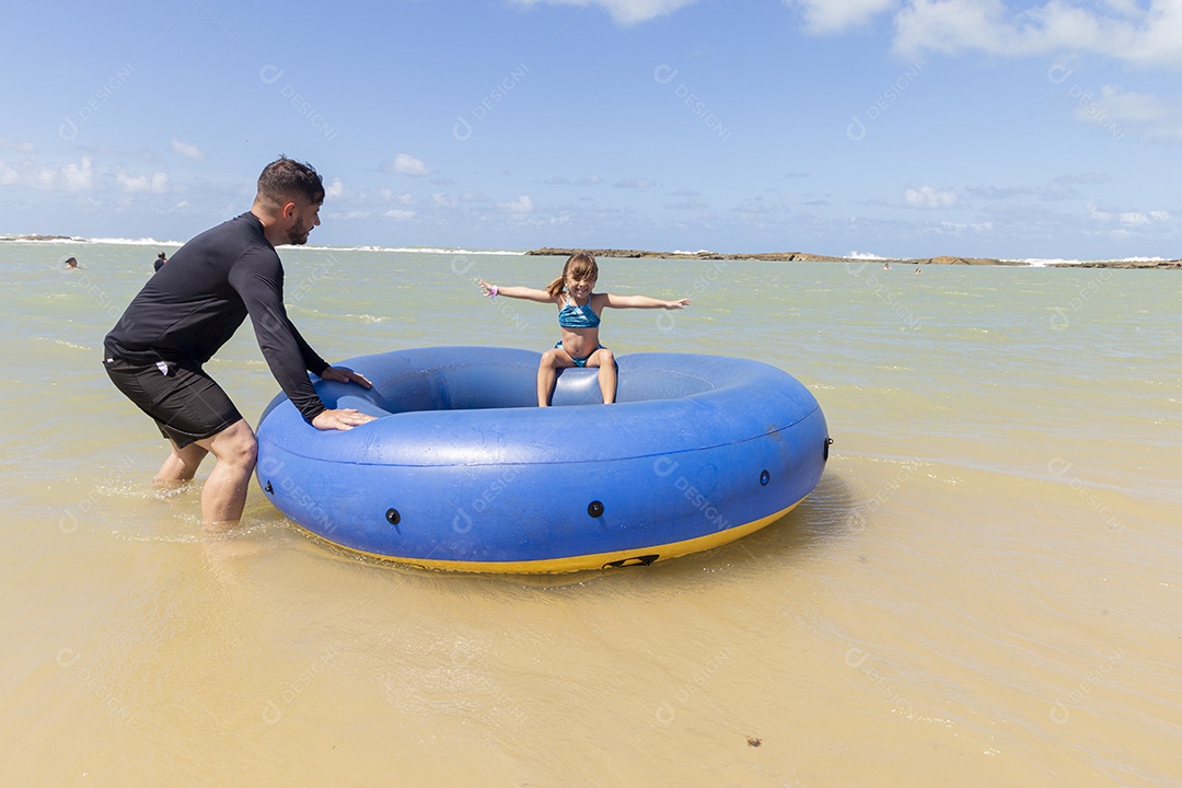 Pai e filha sorridentes brincando com boia azul na praia