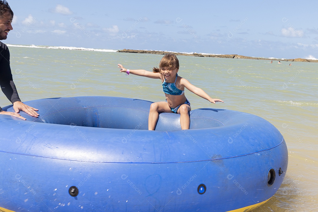 Pai e filha sorridentes brincando com boia azul na praia