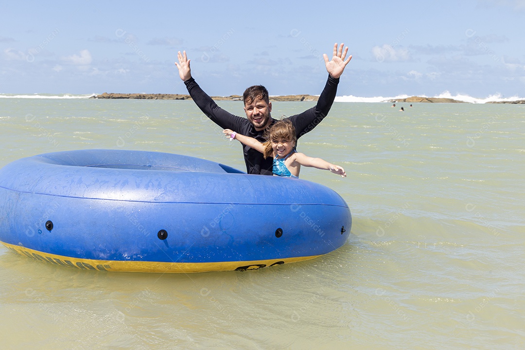 Pai e filha sorridentes brincando com boia azul na praia