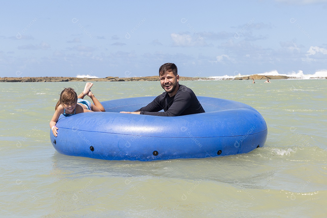Pai e filha sorridentes brincando com boia azul na praia