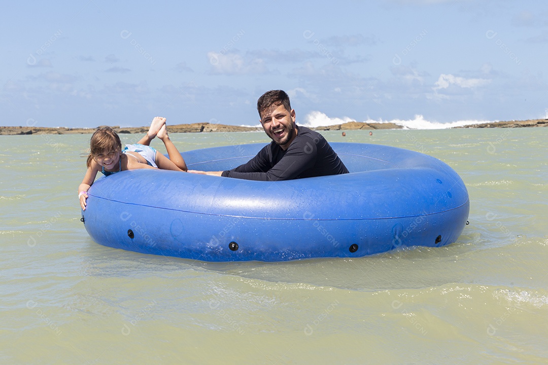 Pai e filha sorridentes brincando com boia azul na praia