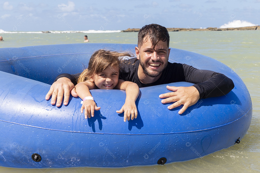 Pai e filha sorridentes brincando com boia azul na praia