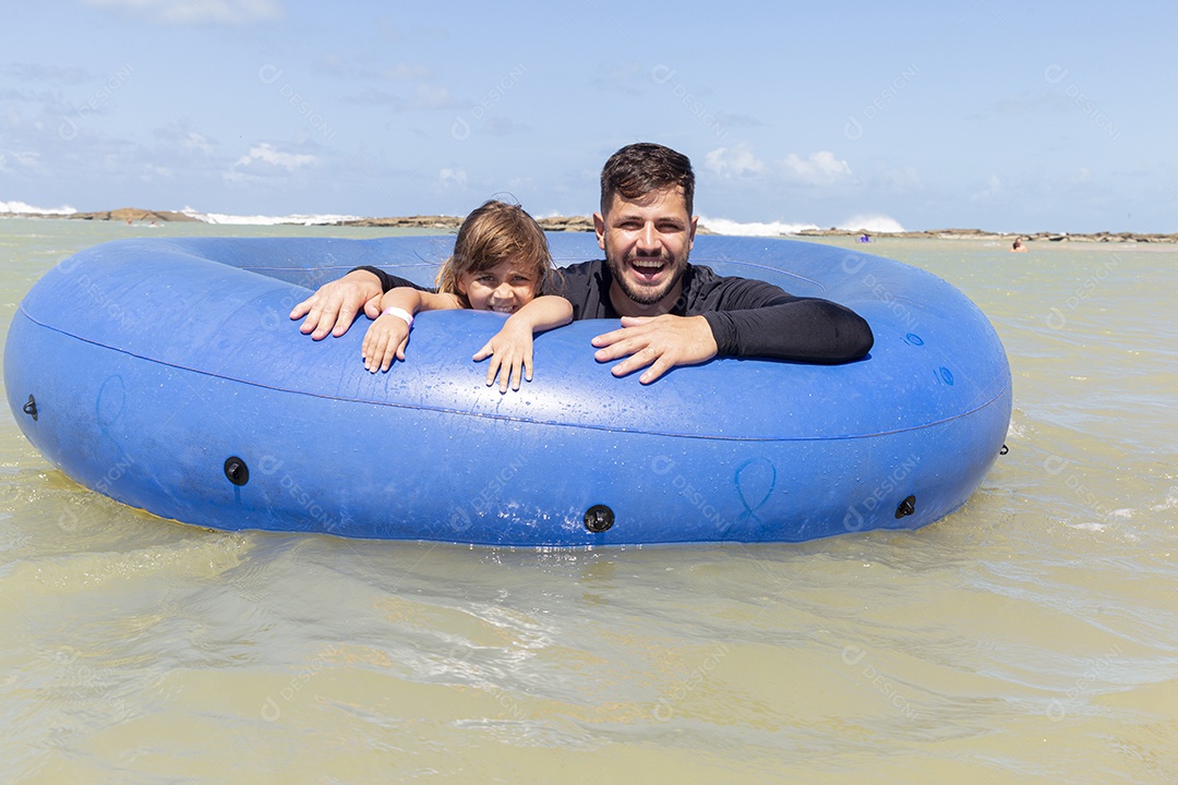 Pai e filha sorridentes brincando com boia azul na praia