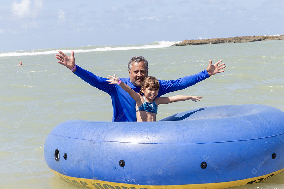 Homem e garotinha sorridentes brincando com boia azul na praia