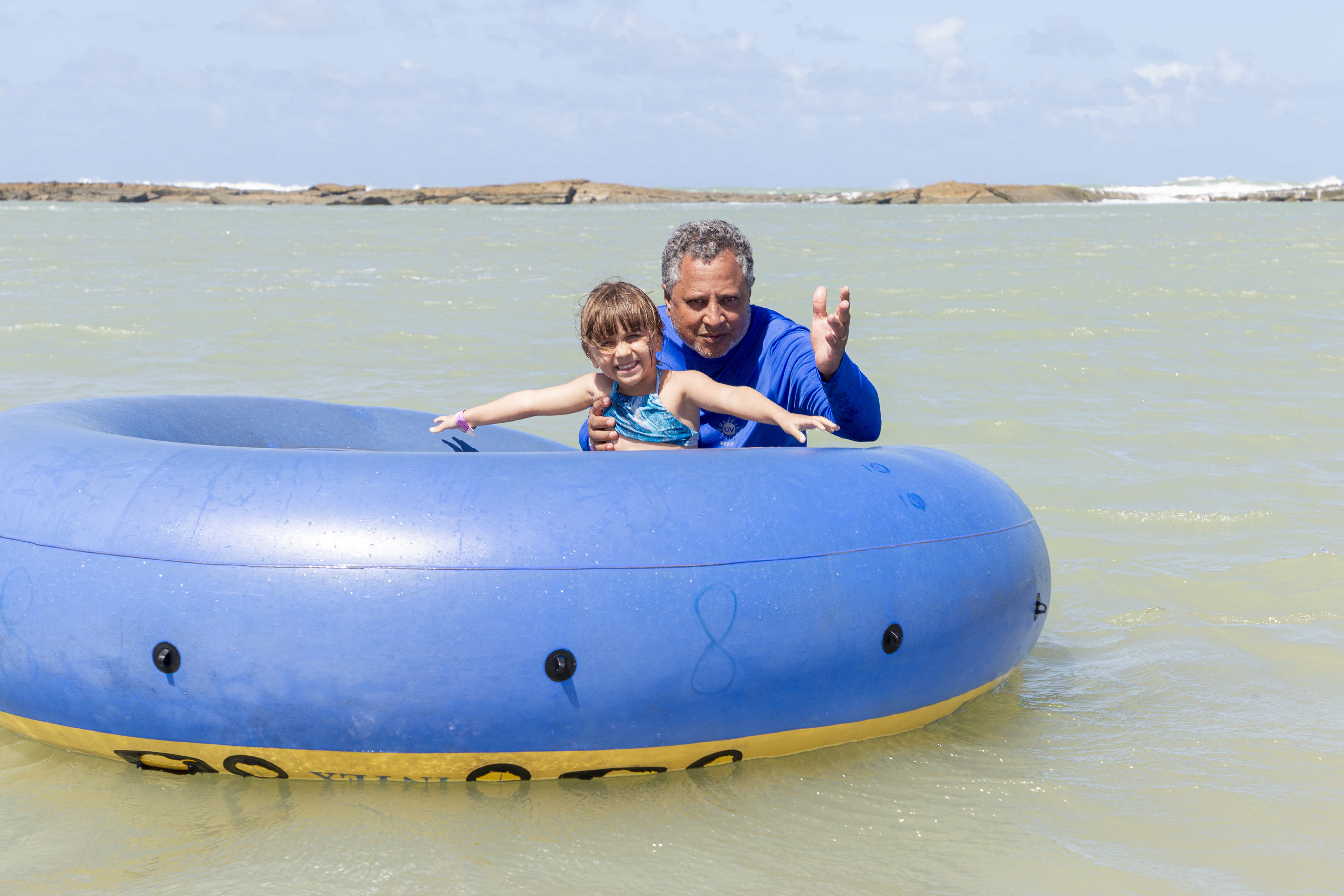 Homem e garotinha sorridentes brincando com boia azul na praia