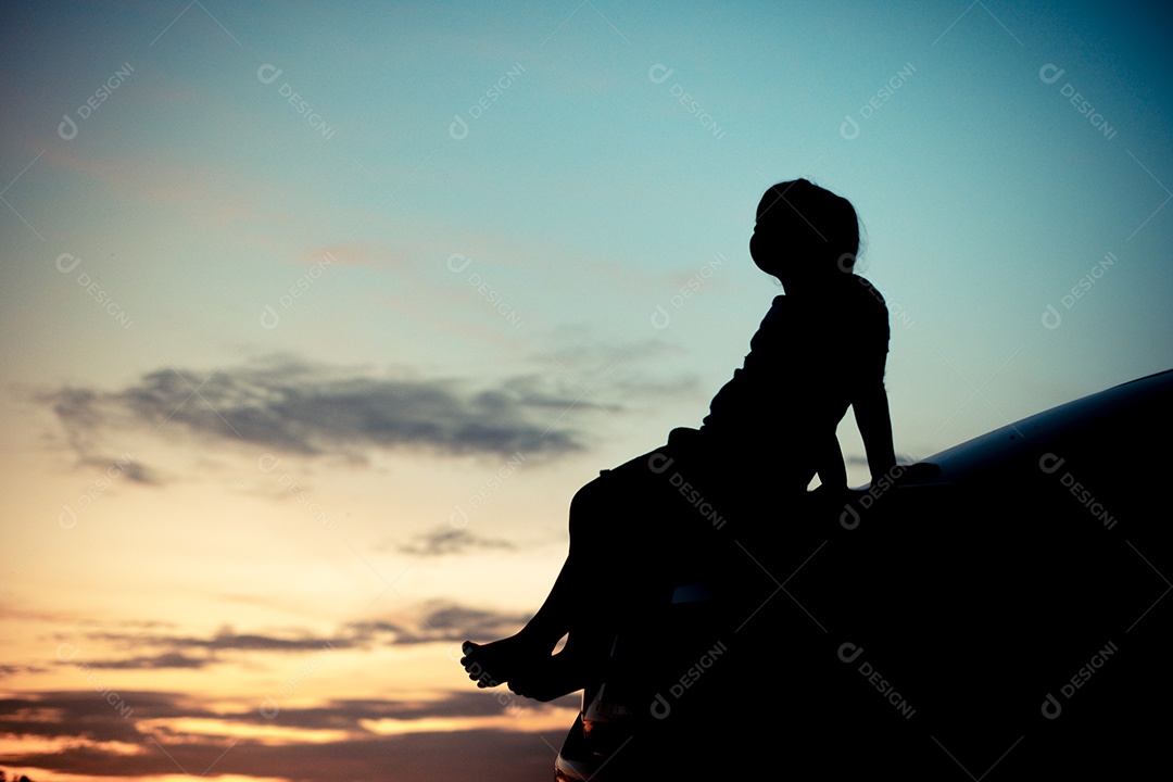 Silhueta de uma garota observando o pôr do sol laranja e o fundo turvo do céu dourado e azul com braços abertos