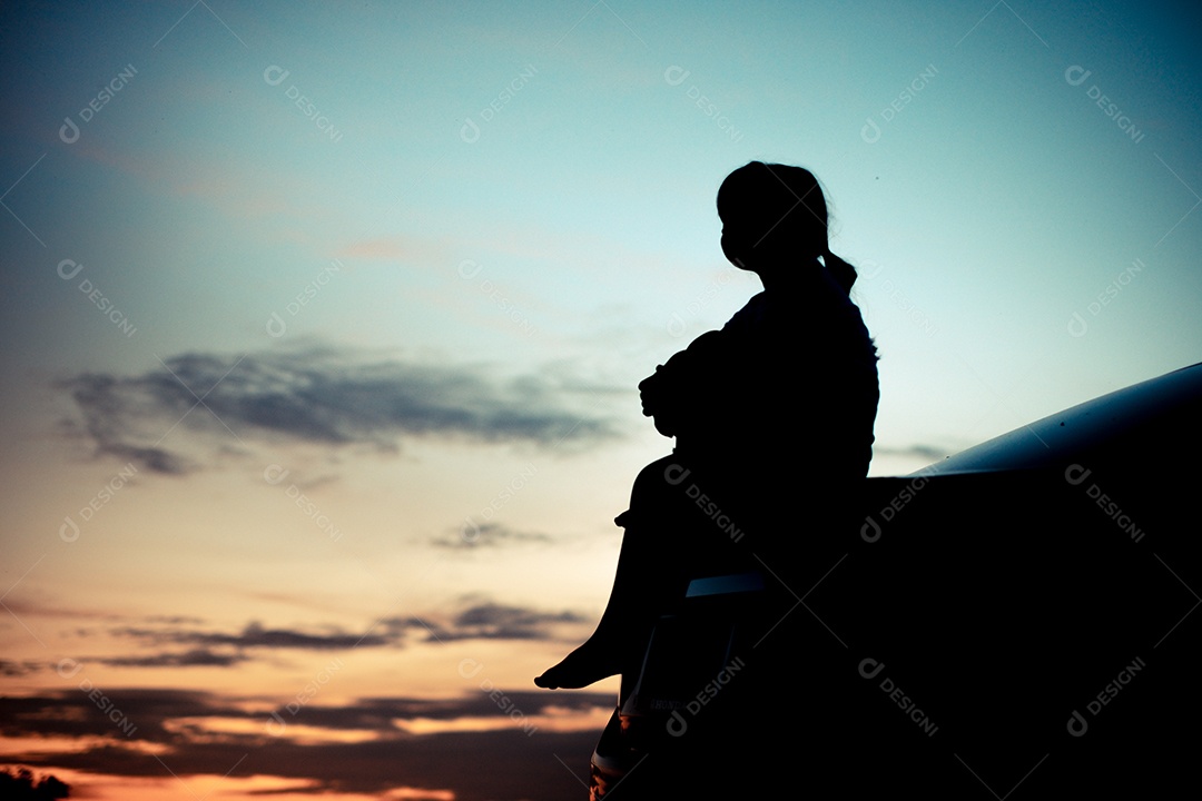 Silhueta de uma garota observando o pôr do sol laranja e o fundo turvo do céu dourado e azul com braços abertos