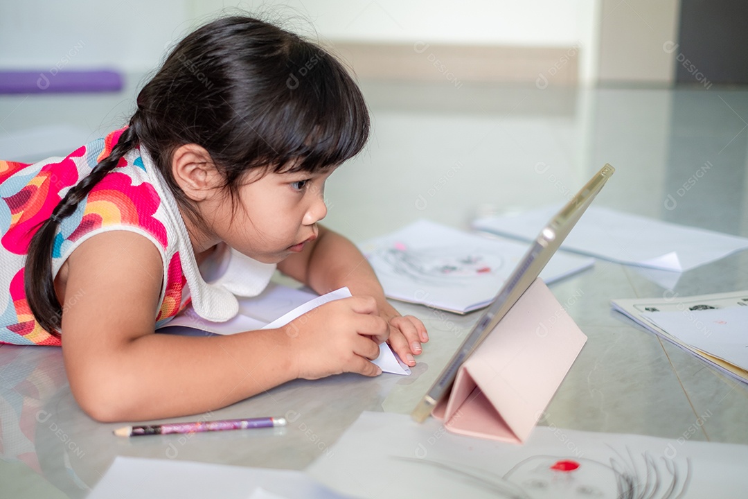 Menina asiática, de 5 a 6 anos, praticando origami usando videoclipes ou estudando on-line via tablet em casa