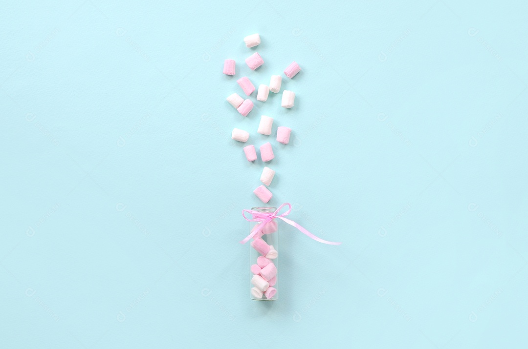 Pedaços de marshmallow rosa e branco são pulverizados de um pequeno recipiente de vidro sobre um fundo azul pastel.